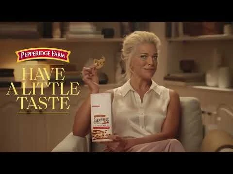 Adult woman holds cookie and package of Pepperidge Farm Farmhouse Thin & Crispy cookies indoors in a light-filled room.