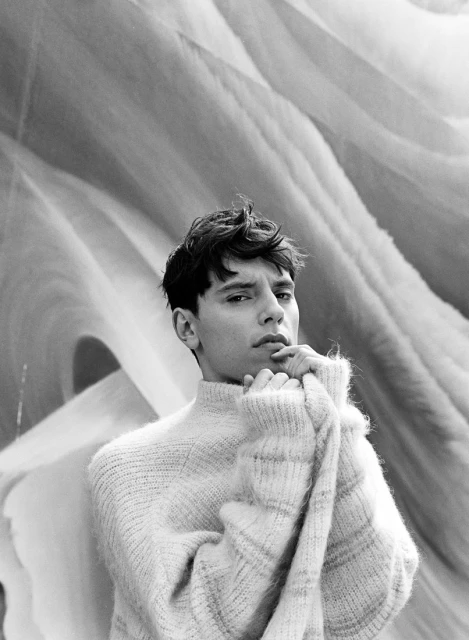 Black and white portrait of a young adult male in a sweater, looking at the camera with a soft expression. Background is textured.