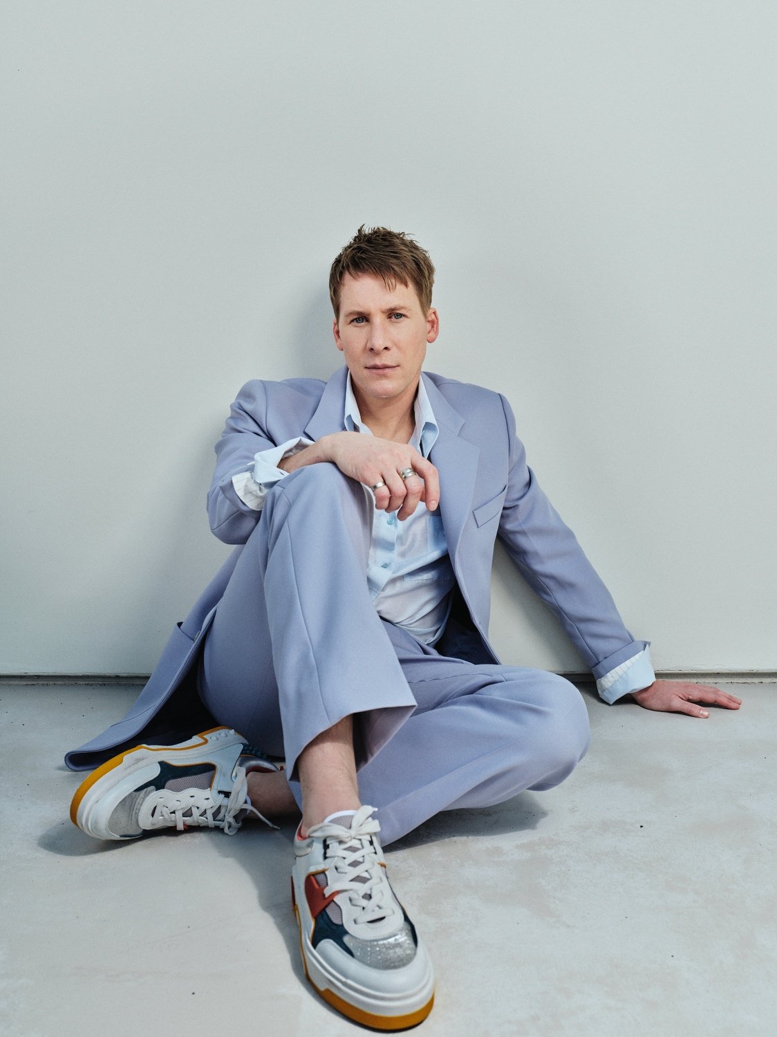 A young adult man in a light blue suit and sneakers is sitting on the floor against a plain wall, posing for a fashion portrait.