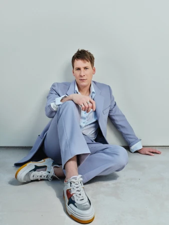 A young adult man in a light blue suit and sneakers is sitting on the floor against a plain wall, posing for a fashion portrait.