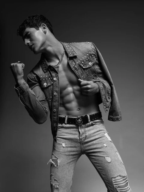 Male model poses in a studded denim jacket and ripped jeans against a gray background, showcasing his abs and muscular physique.
