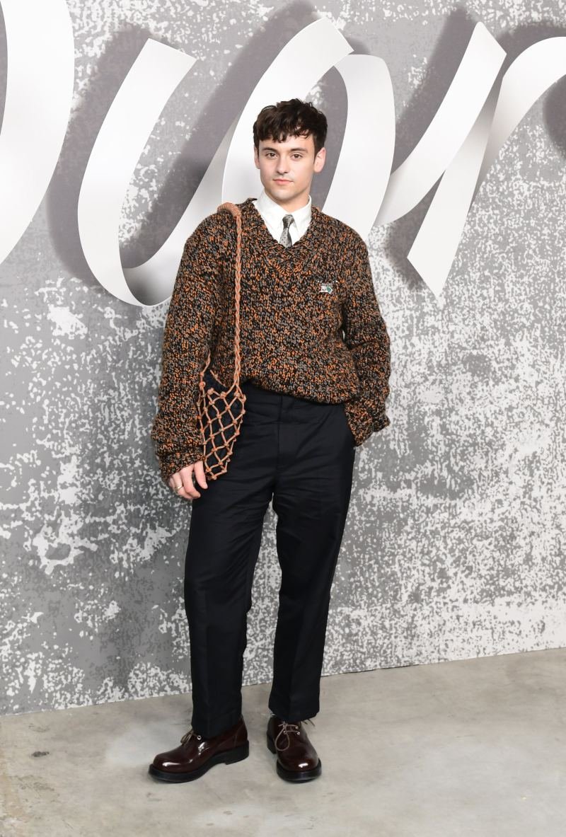 A person poses in a patterned sweater, black pants and brown loafers in front of a logo wall for a fashion editorial shoot.