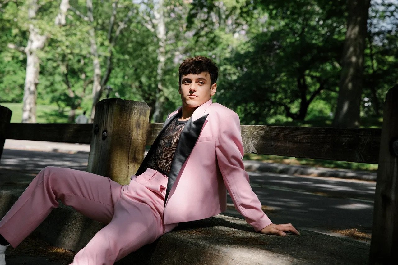 Adult male wearing a pink suit poses on a stone structure in a sun-drenched green park with trees and wooden fence in the background.