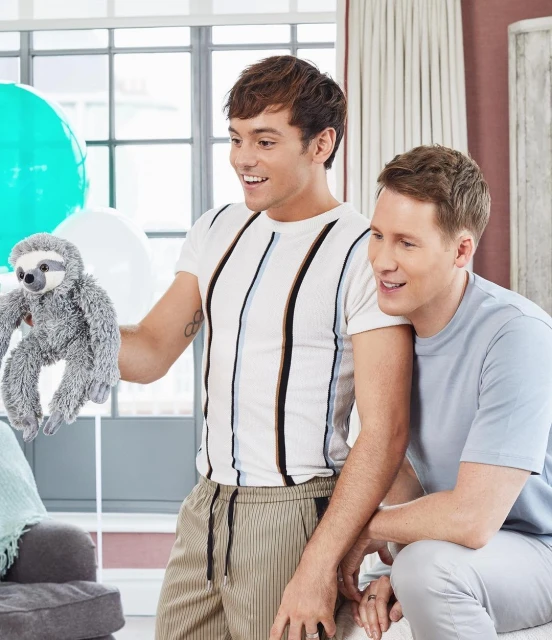 Two adult males, one holding a stuffed sloth toy, are indoors near a window and curtains, with balloons visible, under natural light.