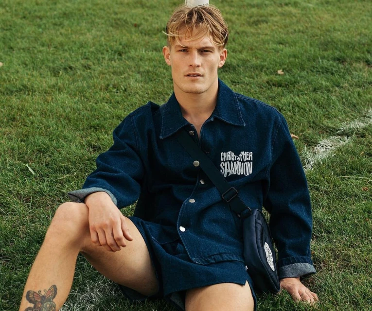 Young adult male sits on grass wearing a blue denim outfit and crossbody bag, looking at the camera on a sunny day.