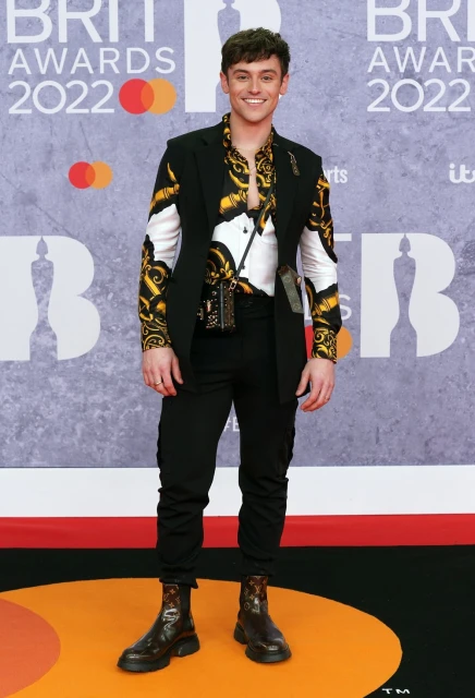 Full-length shot of smiling man in patterned shirt, blazer, pants, and boots at the BRIT Awards 2022 with event branding in background.