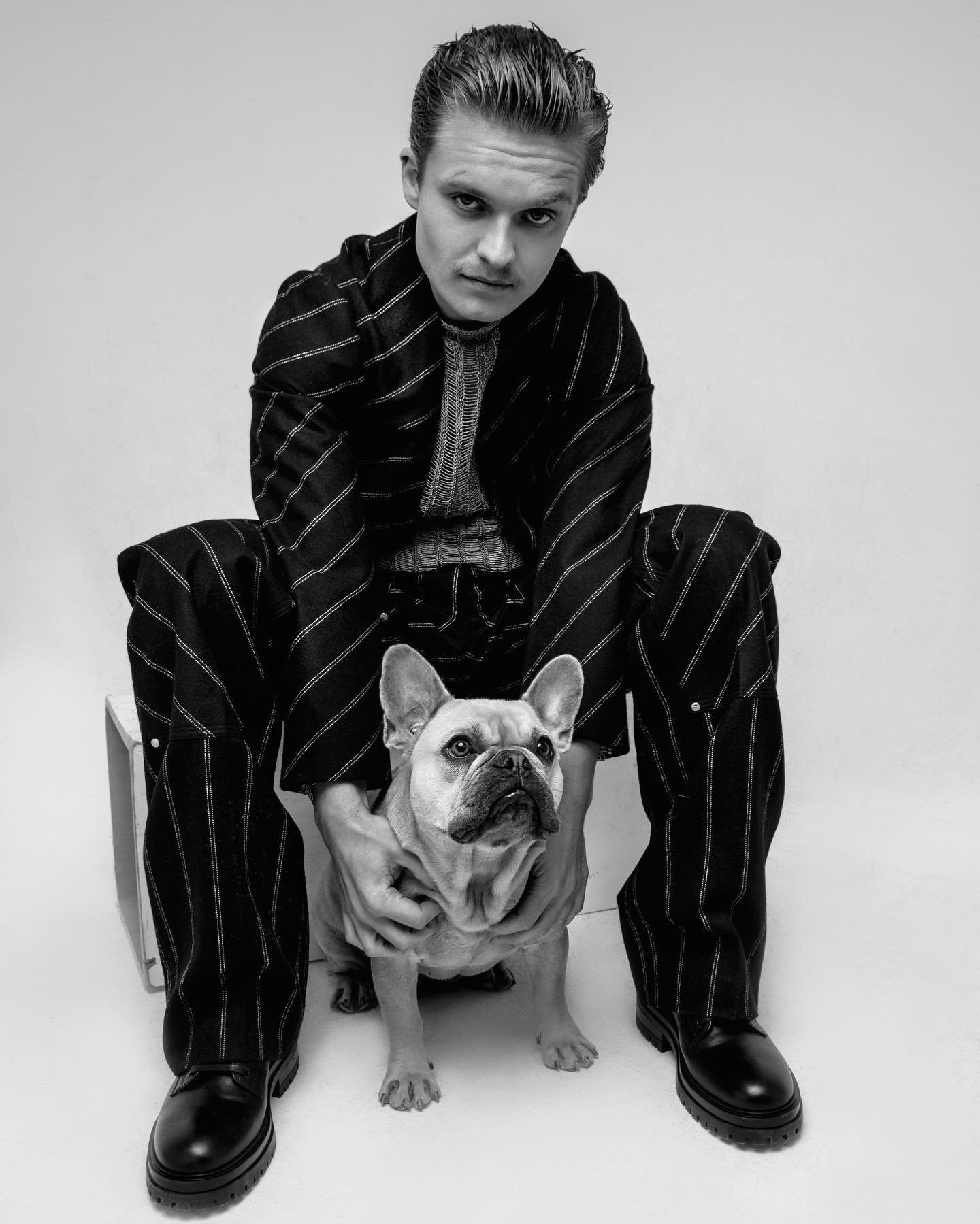 Portrait of an adult male in a striped suit sitting with a French bulldog, captured in a black and white photo studio setting.