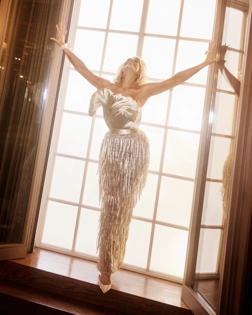 A woman in an elegant dress with fringe details stands near a window, arms raised in a joyful pose, bathed in soft, diffused light.