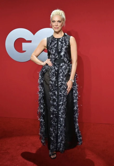 Blonde woman in a black and white fringe gown on the red carpet in front of a red background and the silver GQ logo.