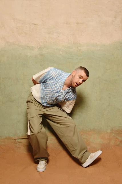 Full-length portrait of a person with short hair leaning back, wearing a patterned button-down shirt, cargo pants, and sneakers, indoors.