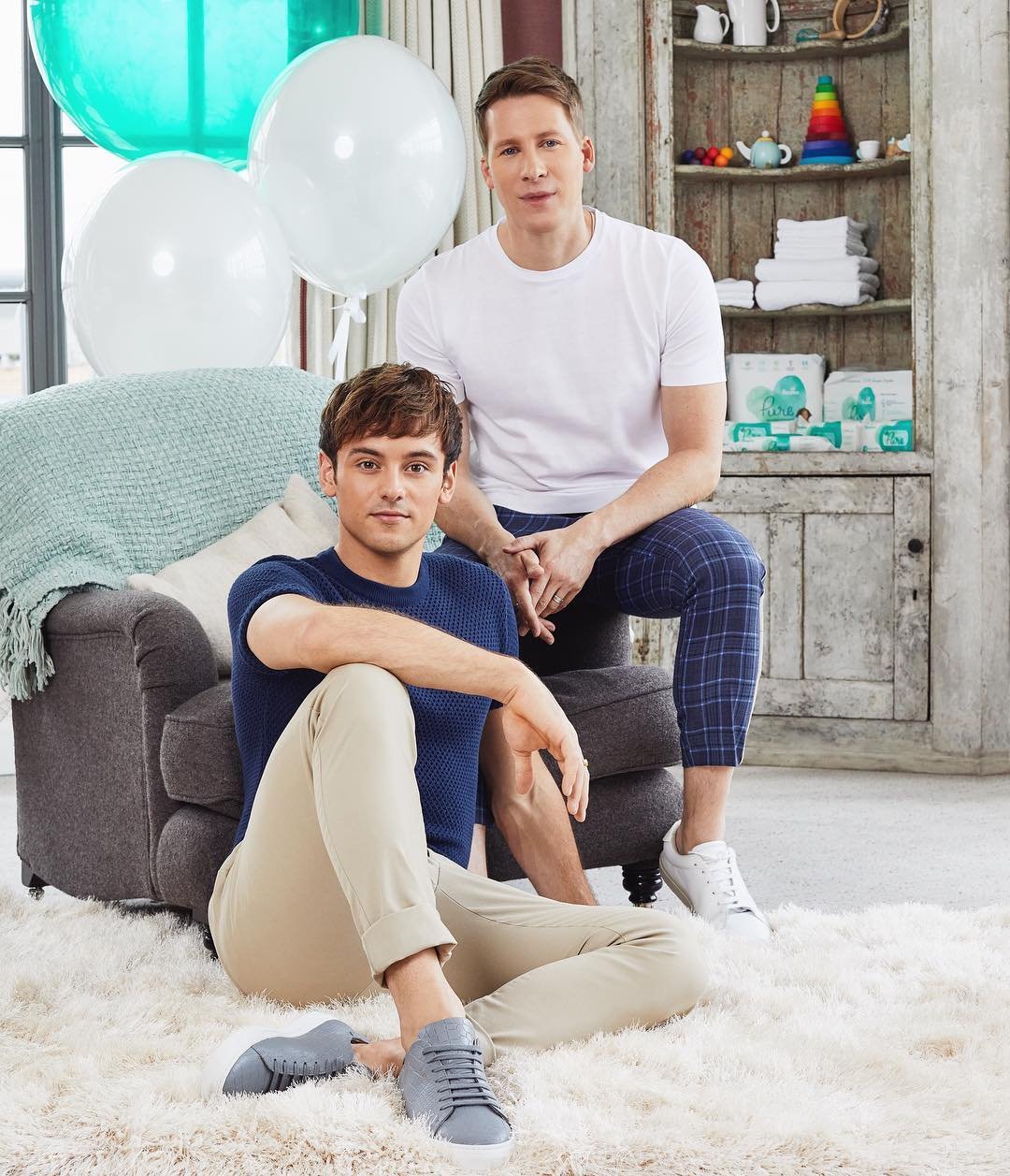 Two adult men pose indoors next to balloons and furniture. One man sits on the floor in beige pants, the other on an ottoman in plaid pants.