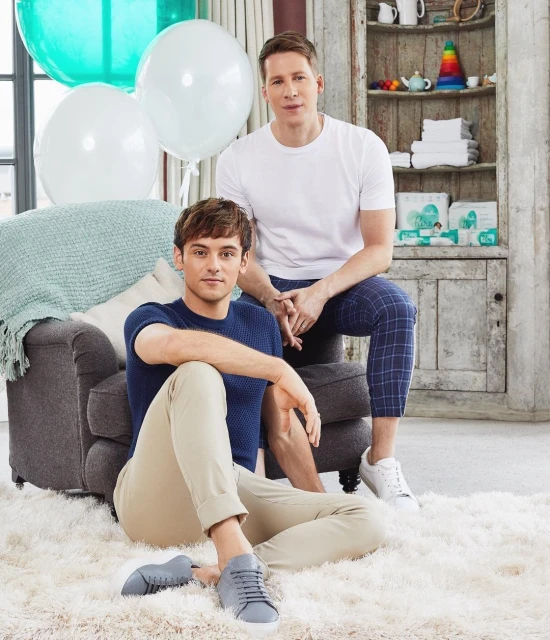 Two adult men pose indoors next to balloons and furniture. One man sits on the floor in beige pants, the other on an ottoman in plaid pants.