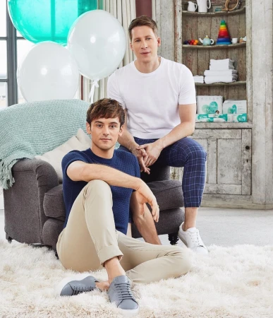 Two adult men pose indoors next to balloons and furniture. One man sits on the floor in beige pants, the other on an ottoman in plaid pants.