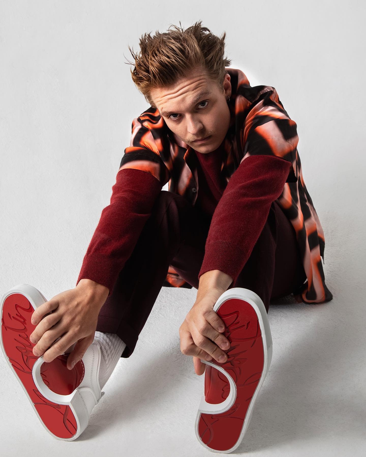 Man sitting indoors wearing a patterned shirt and trousers while showing the soles of his white sneakers with red details.