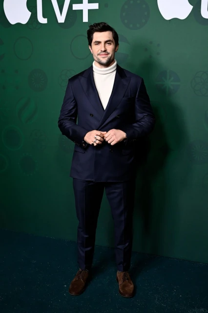 A sharply dressed man in a navy suit and white turtleneck stands against a festive green backdrop with Apple TV+ logo at the top.