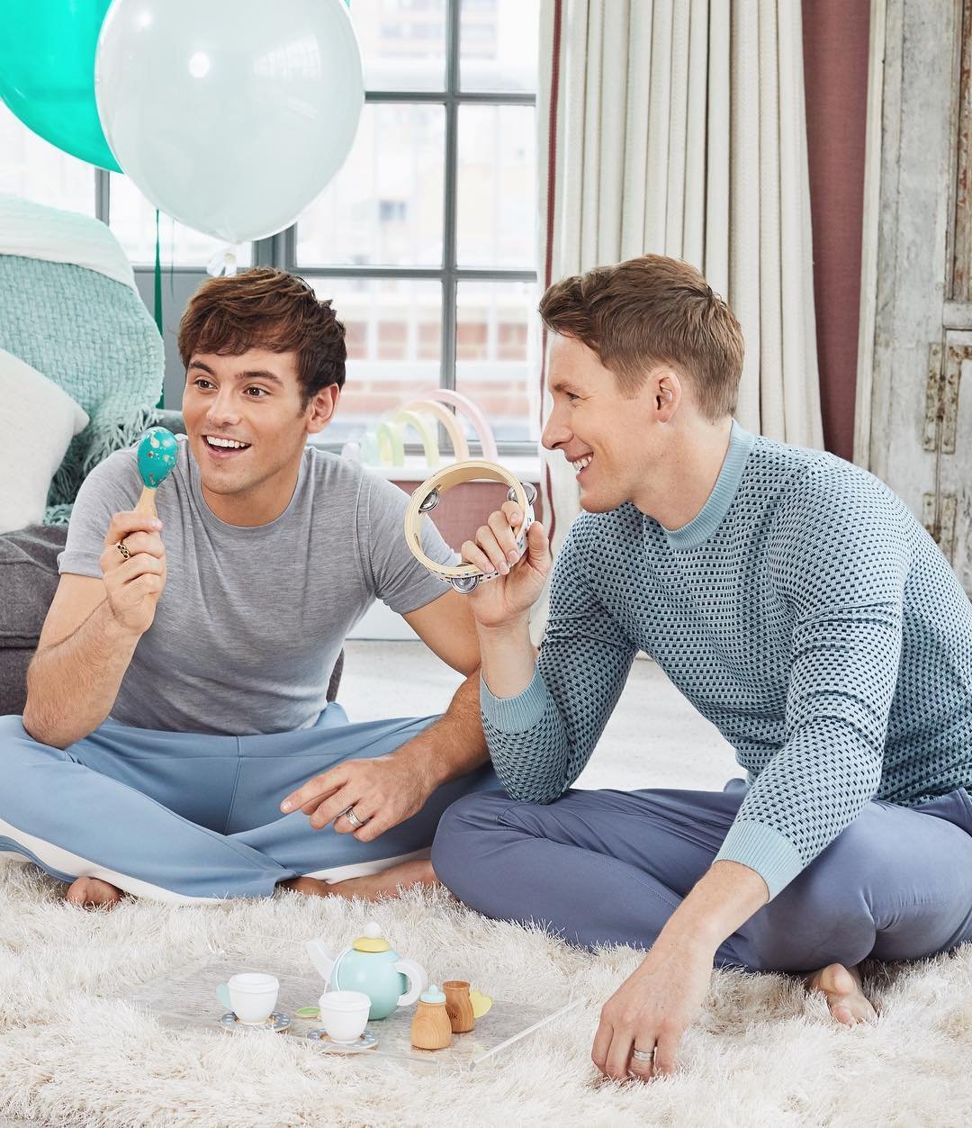 Two adult males sit cross-legged on a white rug, playing with a toy tea set and musical instruments with balloons in the background.