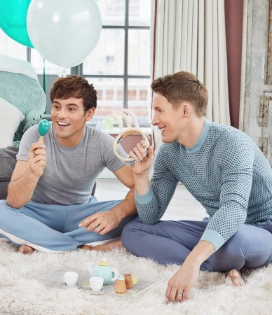 Two adult males sit cross-legged on a white rug, playing with a toy tea set and musical instruments with balloons in the background.