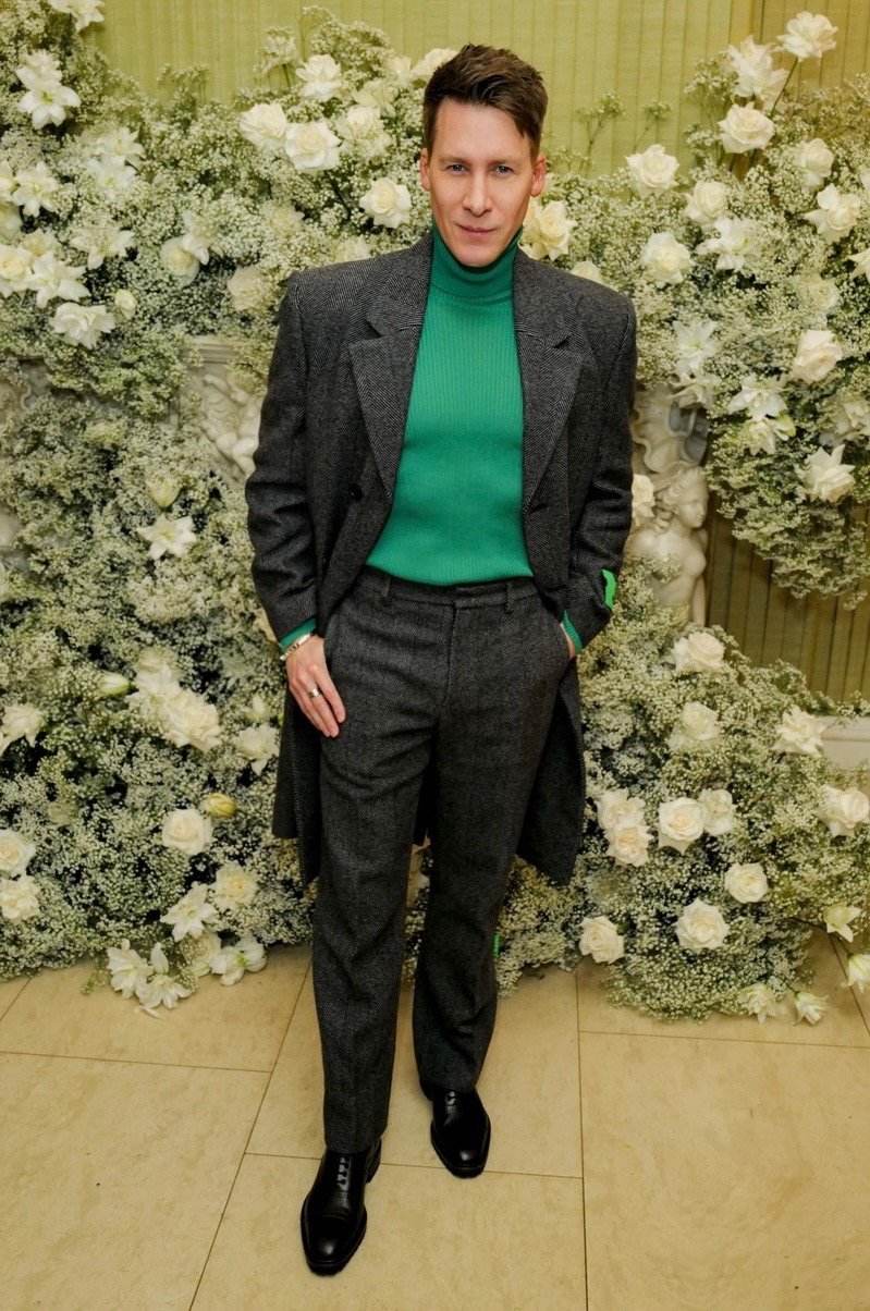 Full-length shot of a fair-skinned adult male wearing a grey coat and pants over a green turtleneck sweater, posing in front of a floral wall.