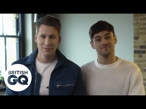 Two adult men pose indoors for a portrait. One wears a blue jacket and the other a white sweater. A 'British GQ' logo is in the corner.