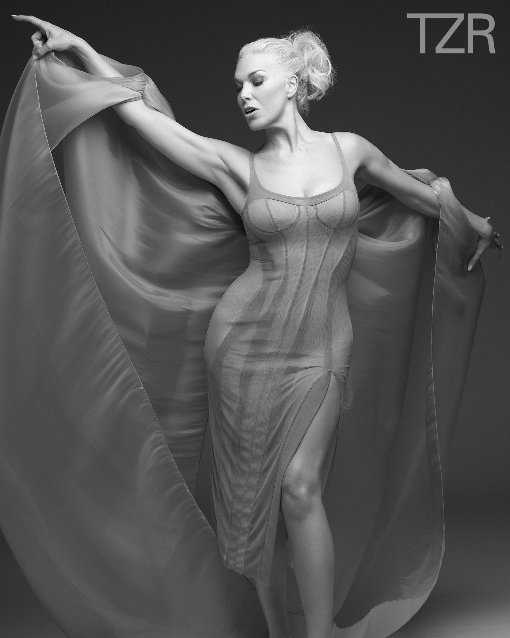 Monochrome portrait of a female model in a flowing dress with arms outstretched against a dark background, exuding elegance.