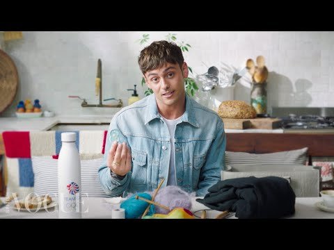 Tom Daley, a brunette male, sits at a table crafting with yarn and knitting needles in a bright indoor setting, wearing a denim jacket.