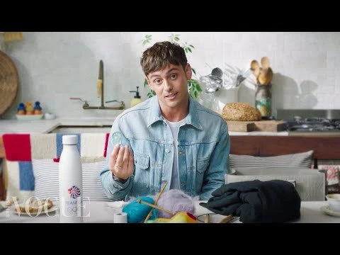 Tom Daley, a brunette male, sits at a table crafting with yarn and knitting needles in a bright indoor setting, wearing a denim jacket.