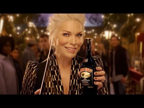 Elegant woman in sequined top holds Baileys bottle in party-like setting with blurred people and lights, promoting a festive mood.