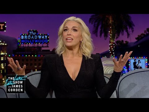 Blonde woman in a black dress sits on a grey chair with a Hollywood sign and palm tree in the background during a late-night TV show.