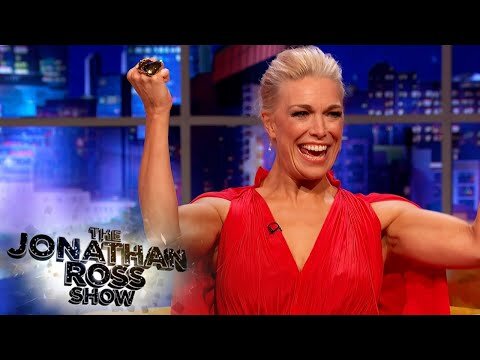 Cheerful adult woman in a red dress raises her arms and smiles on The Jonathan Ross Show set, with an illuminated cityscape background.