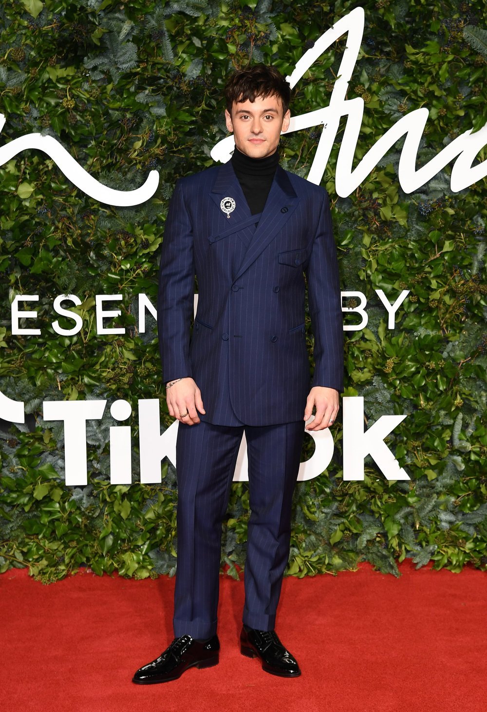 A young adult male in a navy pinstripe suit stands confidently on a red carpet against a leafy backdrop for a fashion event.