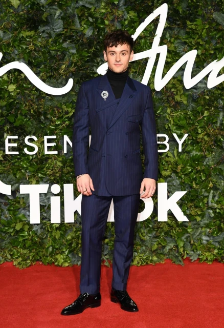 A young adult male in a navy pinstripe suit stands confidently on a red carpet against a leafy backdrop for a fashion event.
