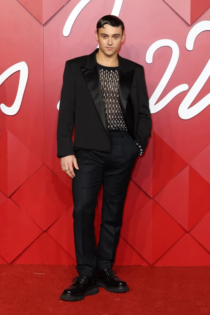 A young adult male in a black suit stands on a red carpet against a red geometric backdrop, looking at the camera in a full shot.