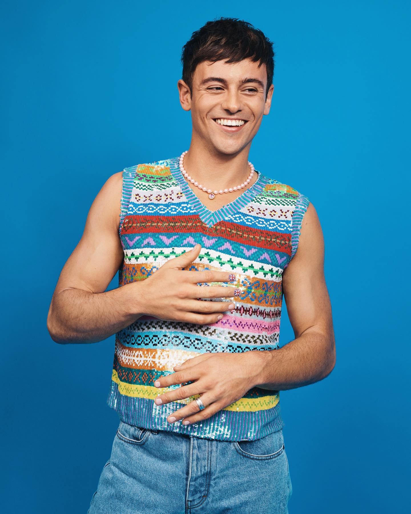 Smiling man in a colorful sweater vest poses against a bright blue backdrop, showcasing a casual yet stylish look with modern flair.