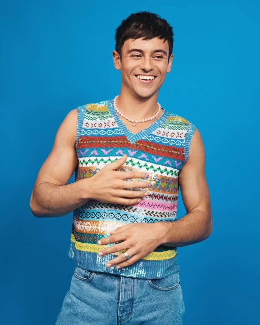 Smiling man in a colorful sweater vest poses against a bright blue backdrop, showcasing a casual yet stylish look with modern flair.