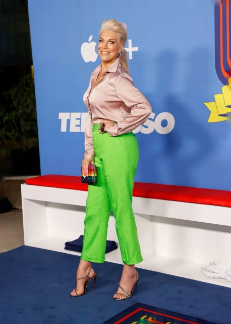 Full shot of a fair-skinned woman with blonde hair, wearing a peach shirt and green pants, standing against a blue backdrop.