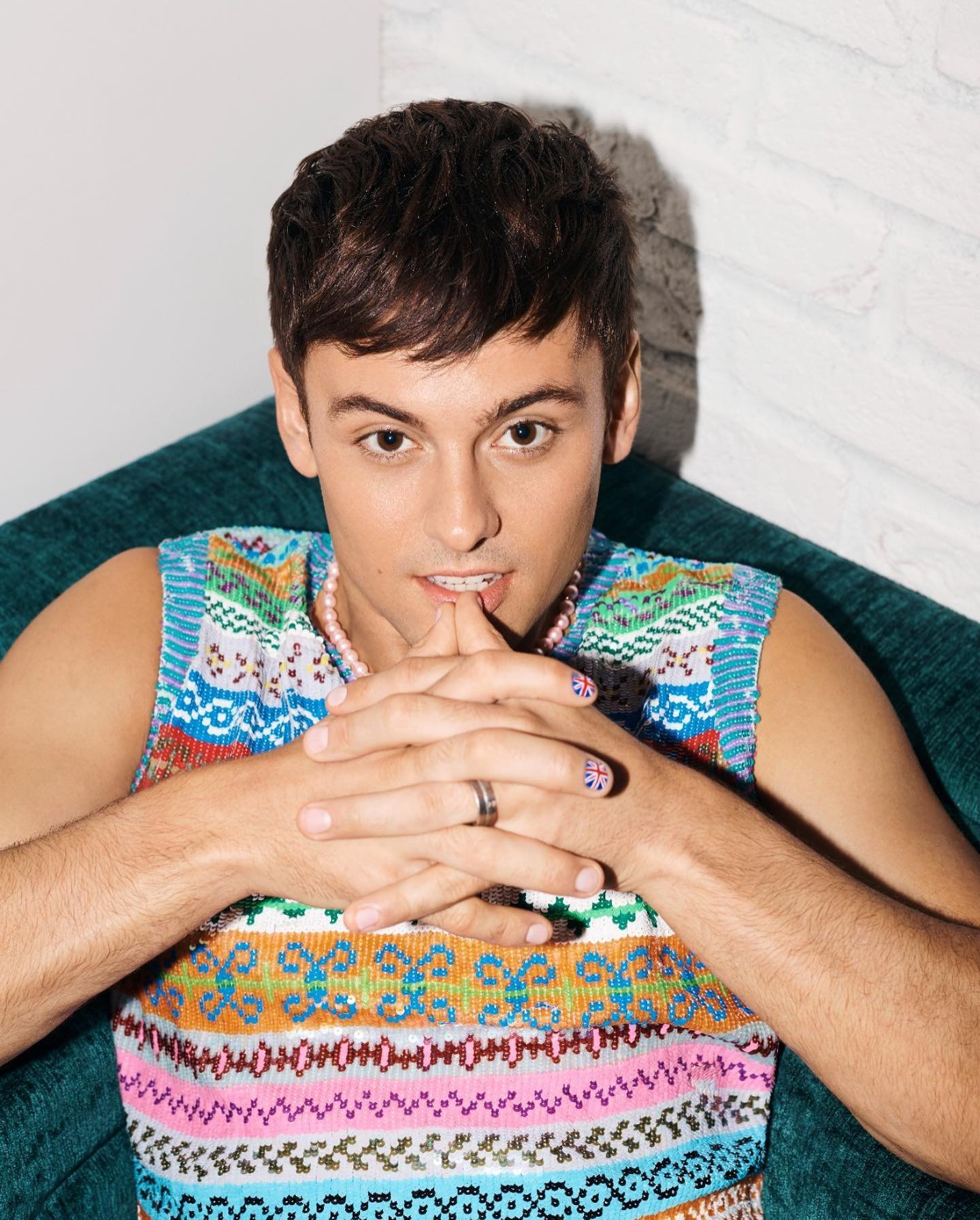 Portrait of a young adult man in a colorful knitted vest with fingers on lips, posing against a white brick wall in a brightly lit studio.