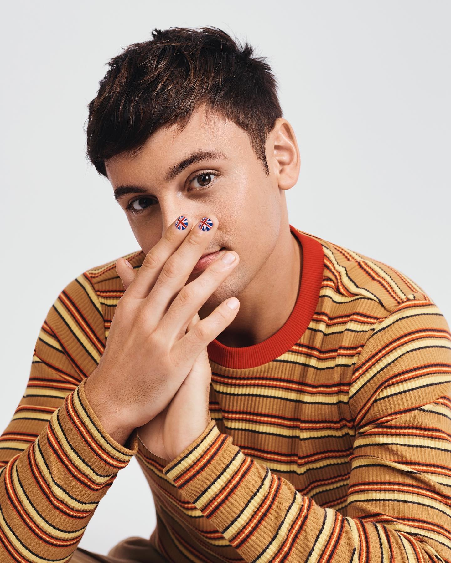 Close-up of a young adult male with painted Union Jack flags on his index fingers, wearing a striped long sleeve shirt in a studio setting.