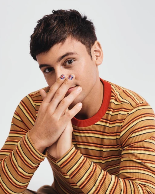 Close-up of a young adult male with painted Union Jack flags on his index fingers, wearing a striped long sleeve shirt in a studio setting.