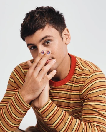 Close-up of a young adult male with painted Union Jack flags on his index fingers, wearing a striped long sleeve shirt in a studio setting.