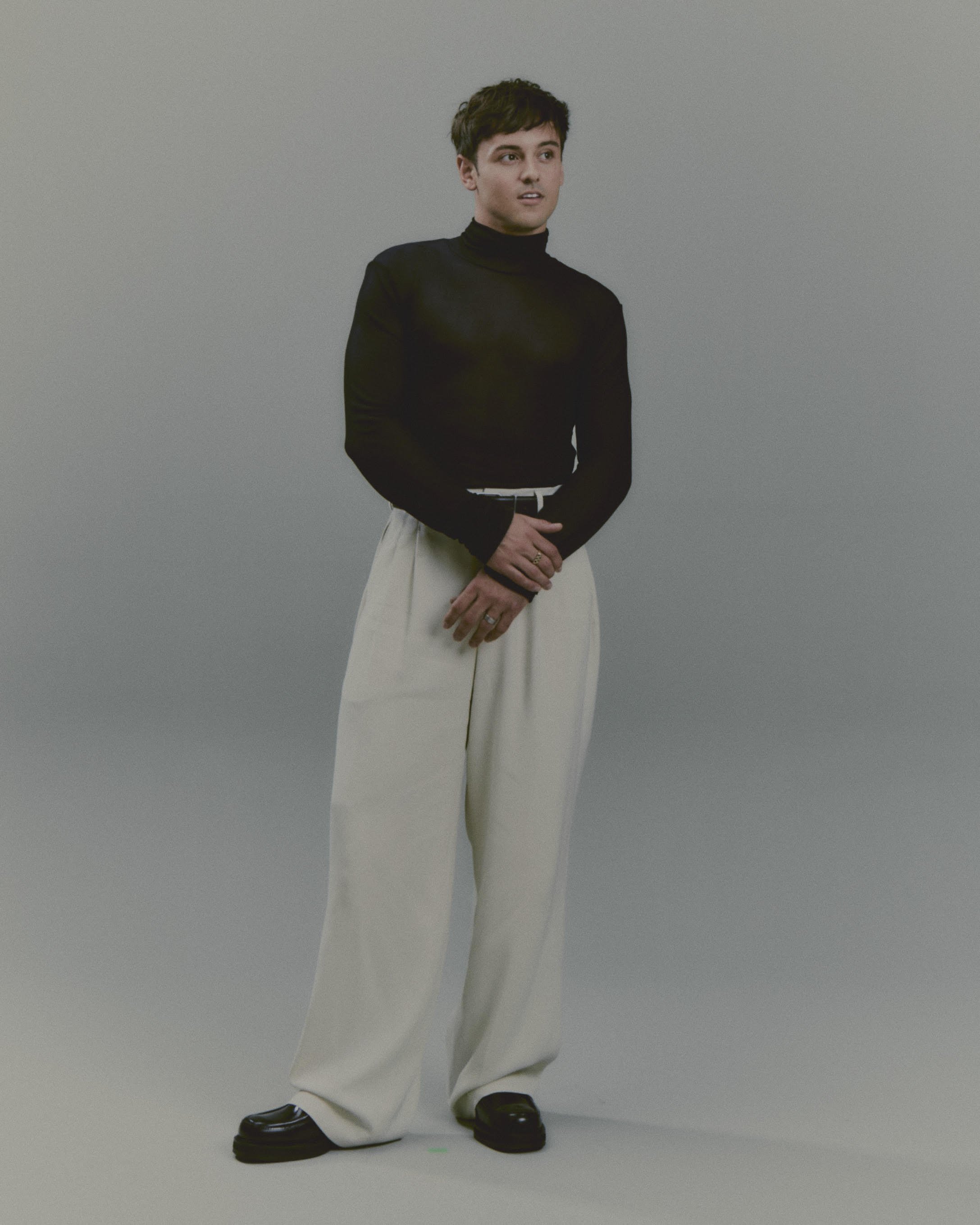 Full-length studio portrait of a man wearing a black turtleneck and light trousers against a neutral background