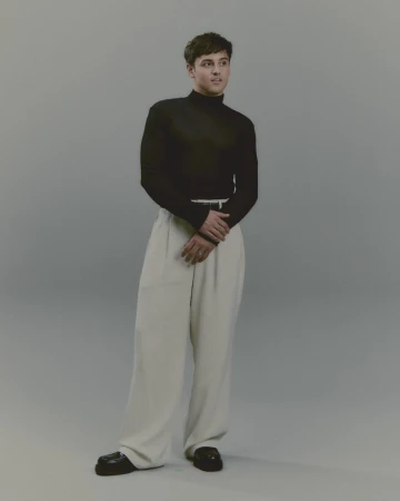 Full-length studio portrait of a man wearing a black turtleneck and light trousers against a neutral background