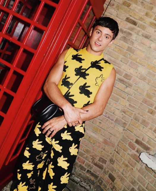 Portrait of a young adult man in a yellow vest and pants with black bunny patterns, posing in front of a red phone booth and brick wall.