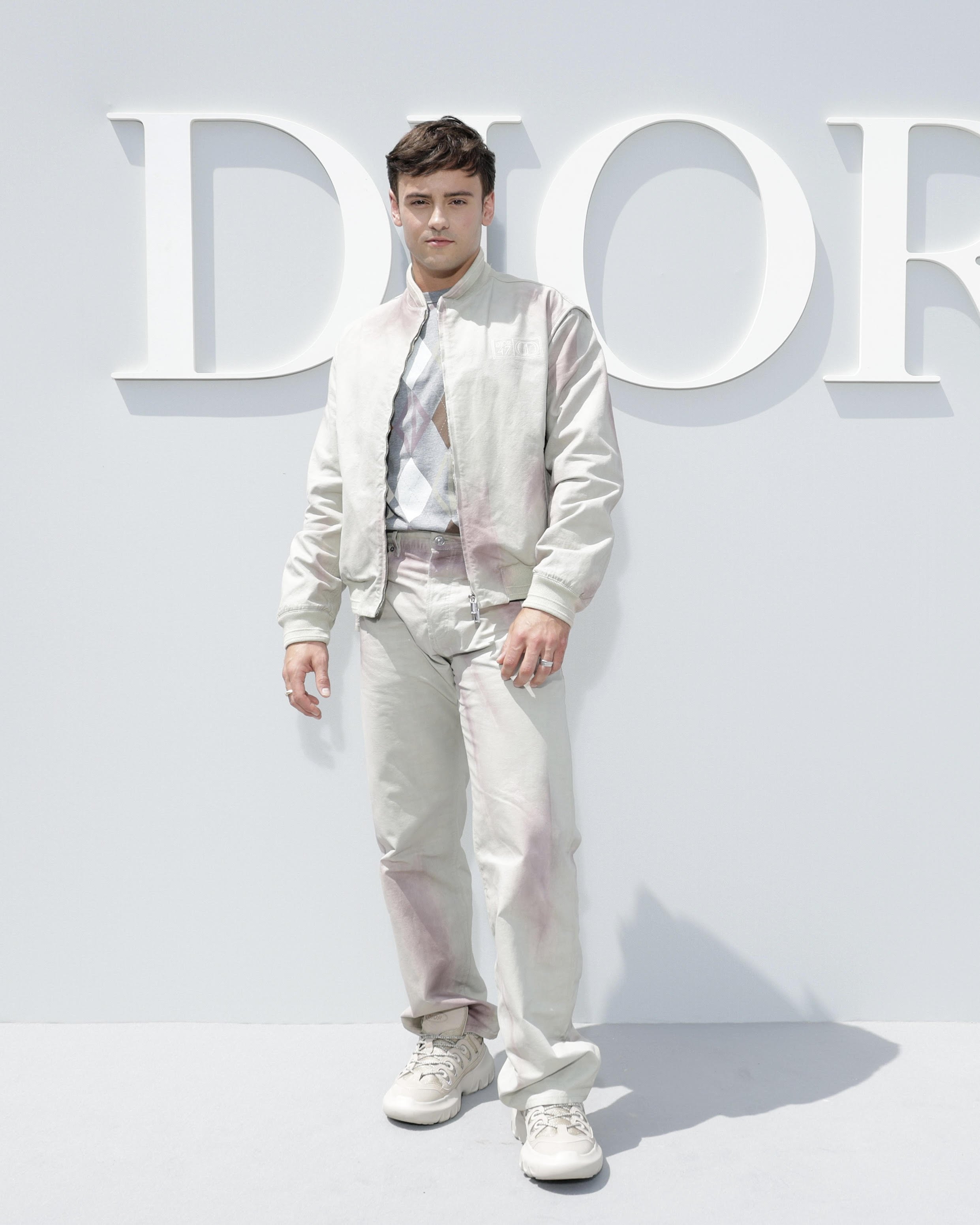 Adult male in a bomber jacket and pants poses in front of a Dior logo. He wears sneakers, and the background is a solid bright white.