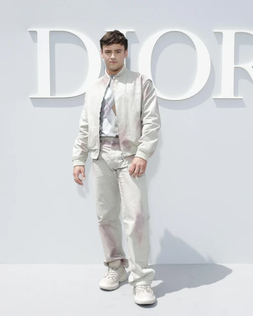 Adult male in a bomber jacket and pants poses in front of a Dior logo. He wears sneakers, and the background is a solid bright white.