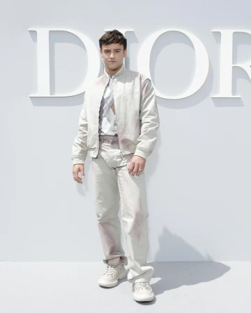 Adult male in a bomber jacket and pants poses in front of a Dior logo. He wears sneakers, and the background is a solid bright white.