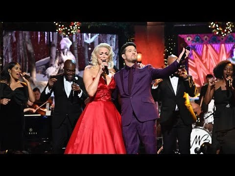 Live performance featuring a male singer in a purple suit, a female singer in a red gown, and a choir singing at microphones on stage.