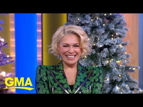 Smiling adult woman with short blonde hair wearing a green patterned jacket, in front of a decorated Christmas tree and backdrop.