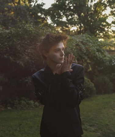 Young man in dark jacket posing outdoors in a garden with hands held near his face styled by James Yardley