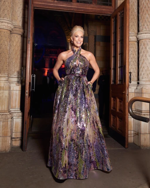 A woman in a formal gown stands in a doorway with a textured stone facade, smiling and looking directly at the camera.