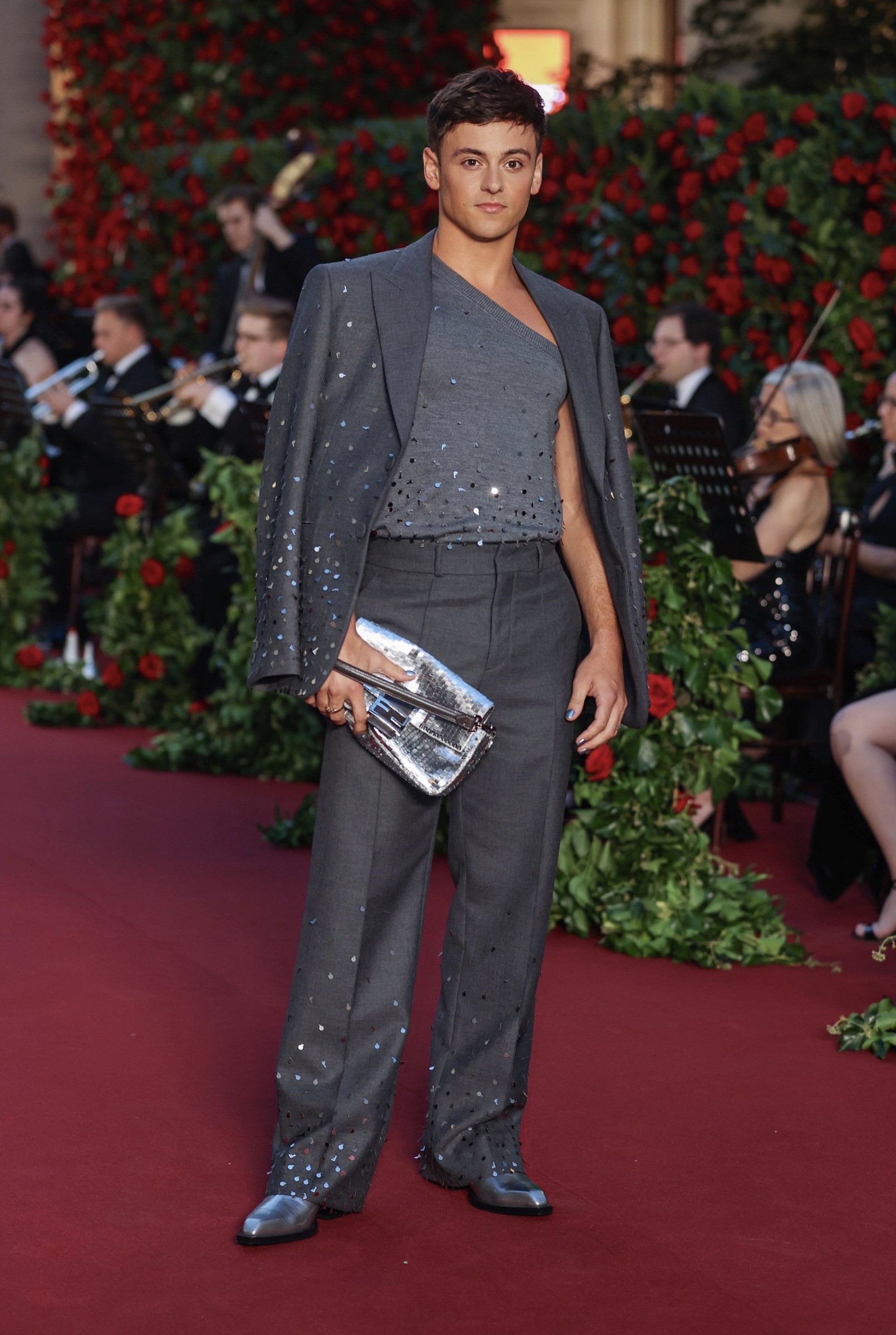 Man in grey suit with silver handbag and shoes walking on red carpet with orchestra and red roses in the background at an event.